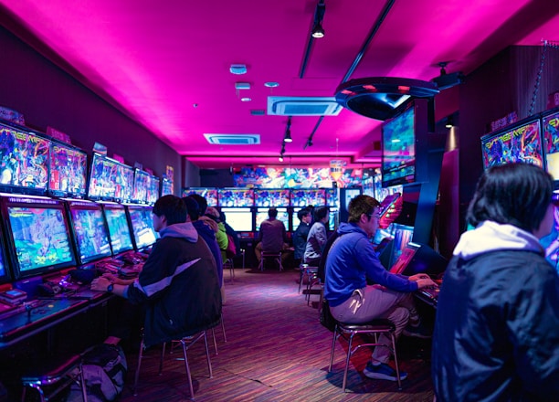 A vibrant arcade with rows of gaming machines adorned with colorful screens. Several people are immersed in playing games, seated on stools in a dimly lit room accented by an intense pink ceiling light. The atmosphere is lively and engaging.