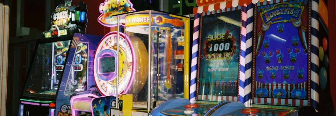 A row of brightly colored arcade game machines is arranged in a line against the wall. Each machine features vibrant lights and designs, with themes ranging from claw games to game show-inspired setups. The area has a playful and inviting atmosphere, suitable for entertainment or amusement centers.