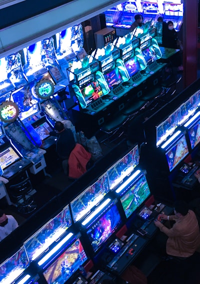 A bustling arcade filled with rows of gaming machines, each with vibrant displays and controls. Several people are seated at these machines, deeply engaged in playing games. Bright fluorescent lights cast a blue and neon glow across the room, enhancing the lively and energetic atmosphere.
